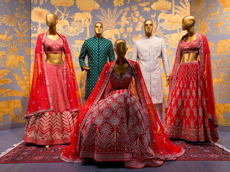 Five gold mannequins in front of a gold and grey background. Three mannequins are wearing red lehengas and two mannequins are wearing green and white kurtas.