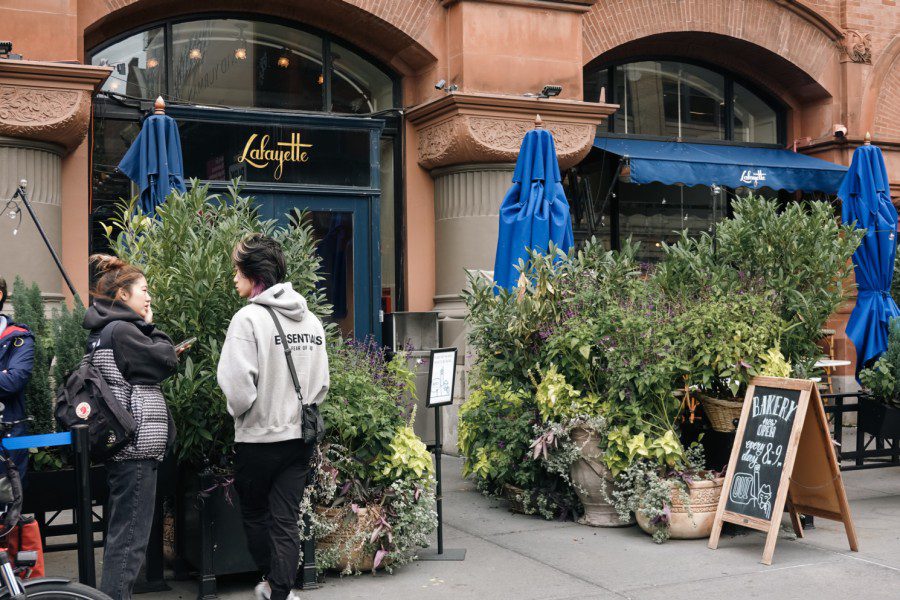 The facade of a restaurant with a lot of plants around the entrance and an outdoor dining area. Next to the plants is a small board that indicates this is a bakery opening from 8 a.m. to 9 p.m. every day.