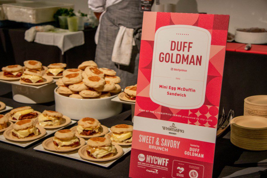 Many English muffin and egg sandwiches stacked in several plates on a black table with a pink sign which reads “DUFF GOLDMAN” and “Mini Egg McDuffin Sandwich,”