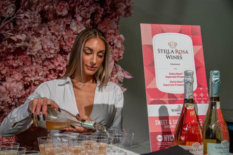 A woman wearing a white shirt pours a bottle of wine into small cups onto a white table, with many pink flowers behind her. Next to her, a pink sign that says “STELLA ROSA WINES.” There are two bottles of wine in front of the sign.