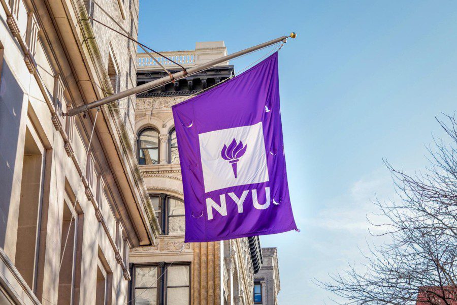 A flag with New York Universitys logo against a purple background hangs down a pole mounted on the exterior of a building.
