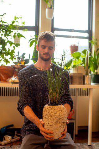 Man sitting in an apartment full of indoor plants, wearing a blue knitted sweater and holding a potted plant which he is looking at.