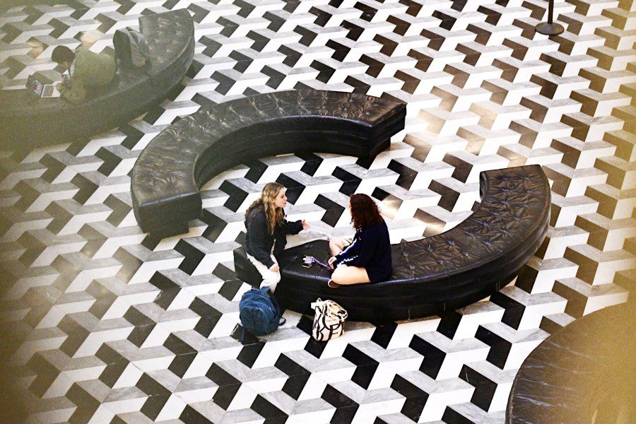 Two people sitting on a circular bench talking to each other with their backpacks on a floor of black-and-white geometrically patterned tiles at Bobst Library.