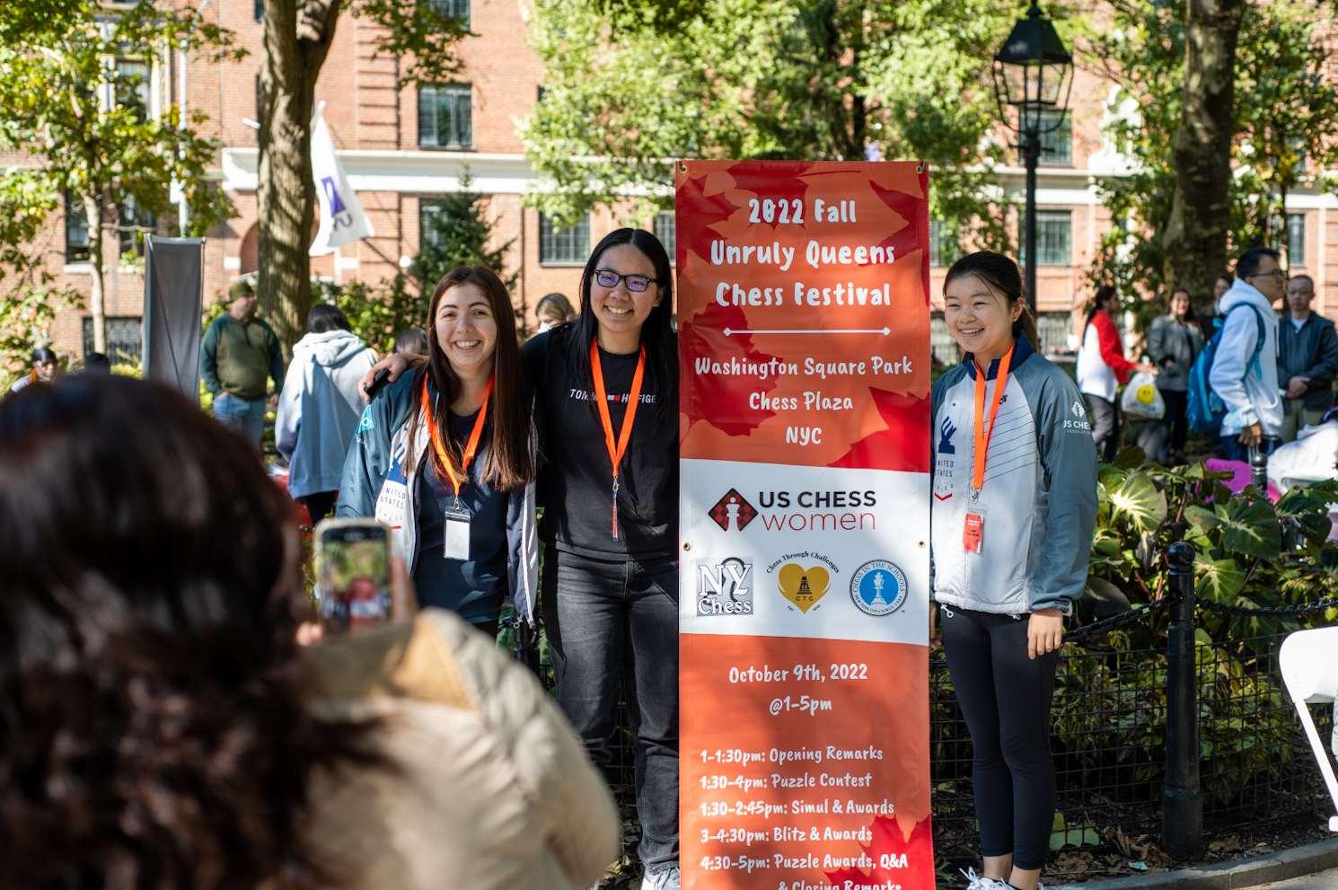 Yassi Ehsani, Evelyn Zhu and Ellen Wang standing by an orange banner that reads “2022 Fall Unruly Queens Chess Festival.”