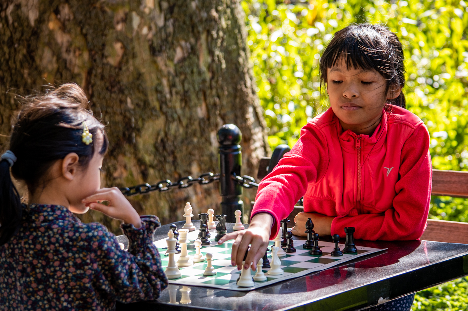 A young girl in a pink fleece holding a piece at a chess board, playing against another young girl.