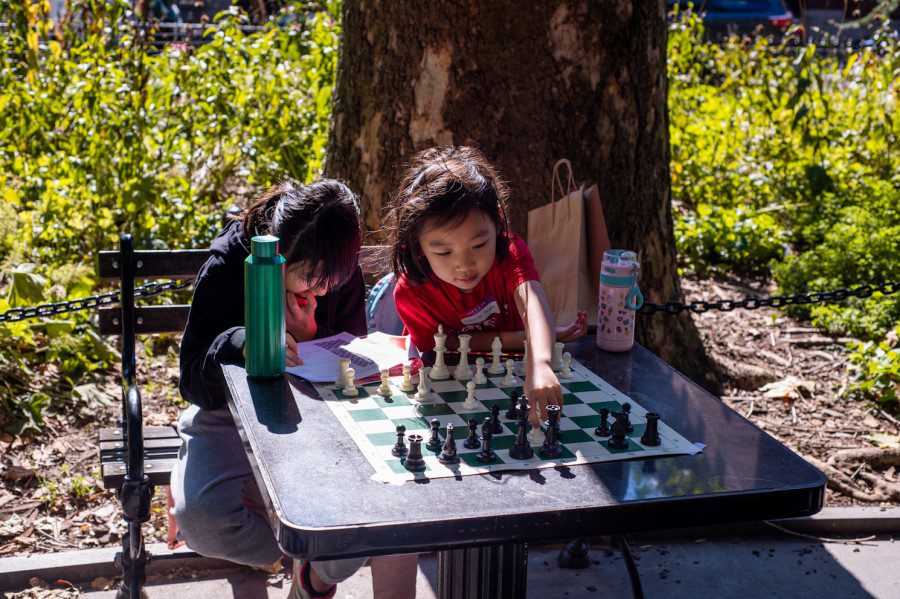 Rui Lin Feng and Olivia Gan sit next to each other at a chess board. Rui Lin Feng is writing in a booklet while Olivia Gan is moving a chess piece.