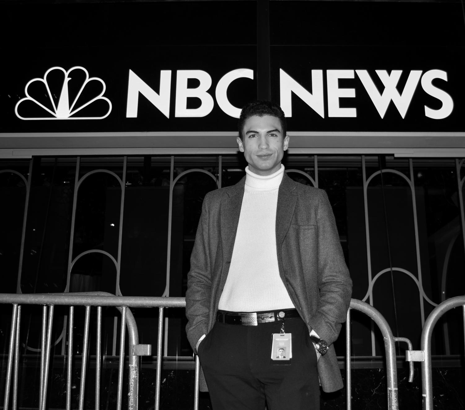 A black-and-white image of a male college student with short dark hair, a light turtle neck, dark pants with keys clipped to the belt loop, and dark blazer standing in front of an N.B.C. News sign. He smiles at the camera.