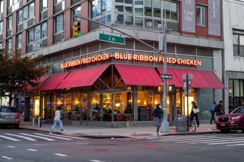 The facade of a restaurant located at a street corner. The storefront has a red awning and red signs which read the name of the restaurant: “BLUE RIBBON FRIED CHICKEN.”