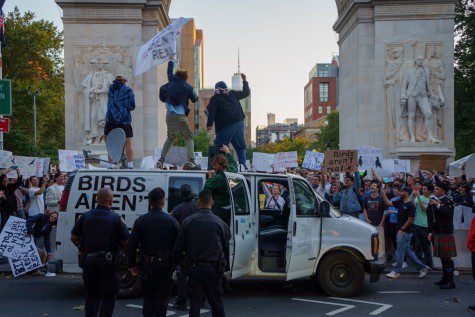 Three people stand on top of a white van which has the text “BIRDS AREN’T REAL” pasted on its side, parked near the Washington Square Arch. A crowd of protestors holds up signs behind the van. Four New York City police officers stand on the other side facing the protesters.