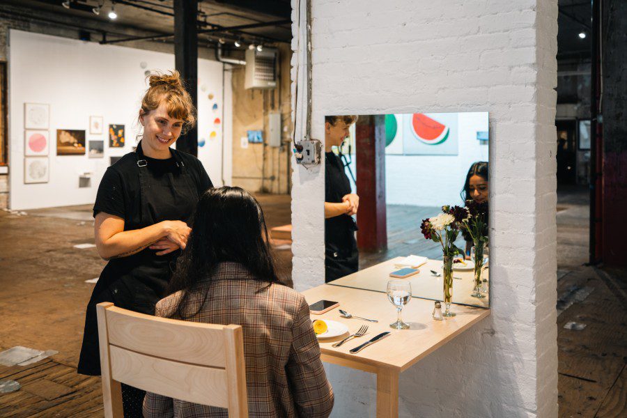 Artist Frida Foberg stands next to her exhibition installation of a wooden table with a mirror, utensils, and dush of plated food on it. WSN contributing writer Alisha Goel sits in a wooden chair by the table facing Frida.
