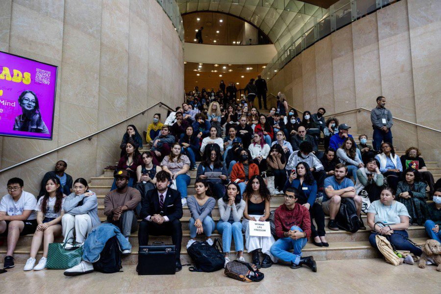 A large group of people sit on the large staircase inside the lobby of New York University’s Kimmel Center for University Life, with one person in the front row holding a sign with text “WOMAN LIFE FREEDOM” printed on it.