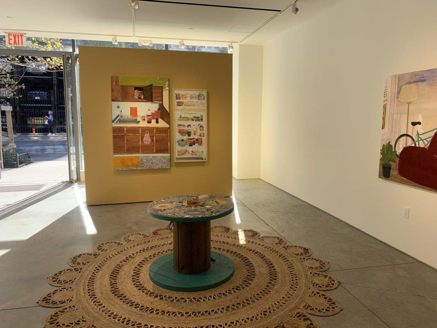 A view of the Praxis Gallery showing a mustard yellow wall with two artworks hanging on it. To the left, a kitchen with snacks and beverages on the countertop. To the right, an art piece depicting an open fridge. To the right of the yellow wall, there is a white wall with a painting of a grey bicycle.