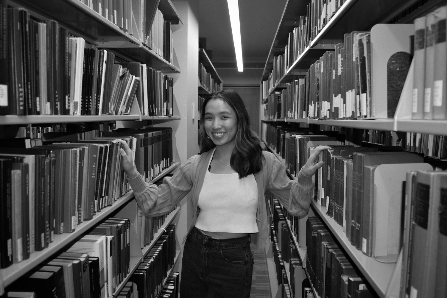 A black-and-white image of a female college student with medium-length slightly wavy dark hair, a light crop top, dark jeans, and a light cardigan smiling at the camera stands in between two bookshelves. Both her hands are placed on books on either side of the shelves.