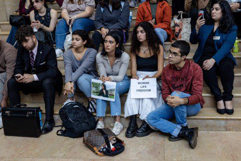 A group of students sitting on the large staircase inside the lobby of New York University’s Kimmel Center for Student Life with one person in the front row holding a sign with text “WOMAN LIFE FREEDOM” printed on it.