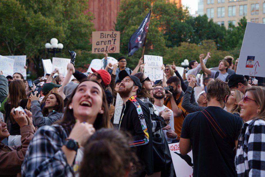 A crowd of protesters at Washington Square Park look up into the sky. Some of them are holding signs with the text “Birds Aren’t Real,” others are making thumbs down gestures to birds flying past.