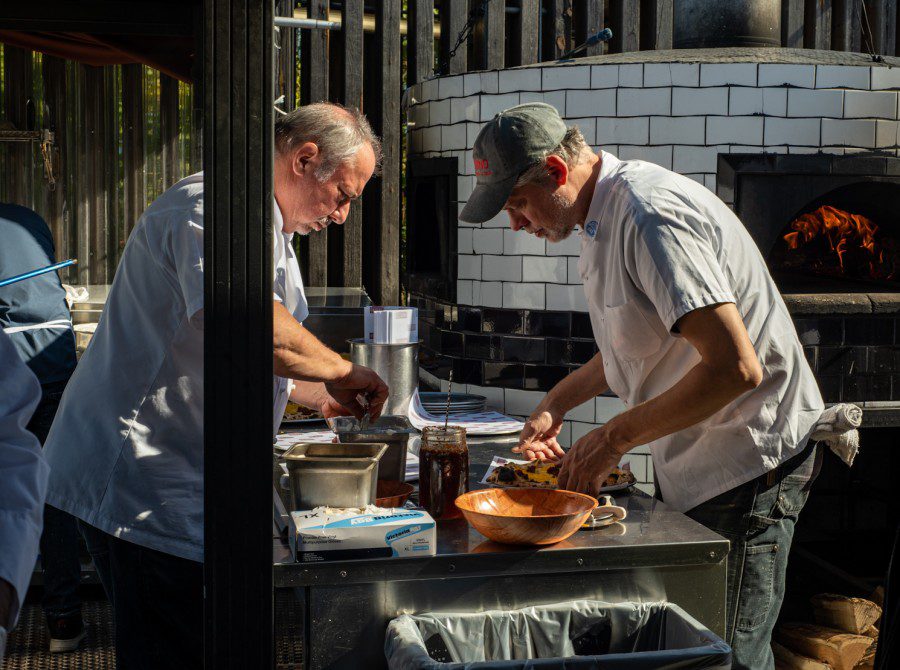 Two chefs in white t-shirts and one in a gray baseball cap add toppings to pizzas.