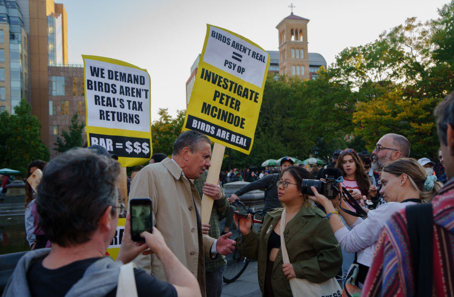 A man in a beige coat speaks to a WSN reporter and a crowd of protesters at Washington Square Park. Behind the man are two people holding up signs that read “WE DEMAND BIRDS AREN’T REAL’S TAX RETURNS,” and “INVESTIGATE PETER MCINDOE. BIRDS ARE REAL.”