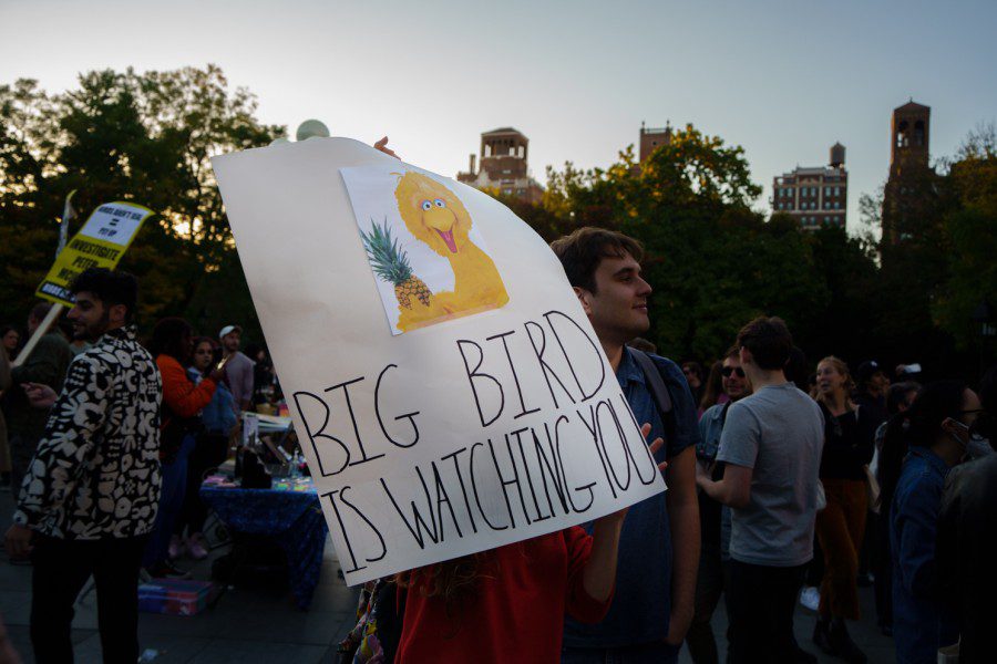 A person wearing a red shirt holds up a white poster with an image of the cartoon character Big Bird and the text “BIG BIRD IS WATCHING YOU.”