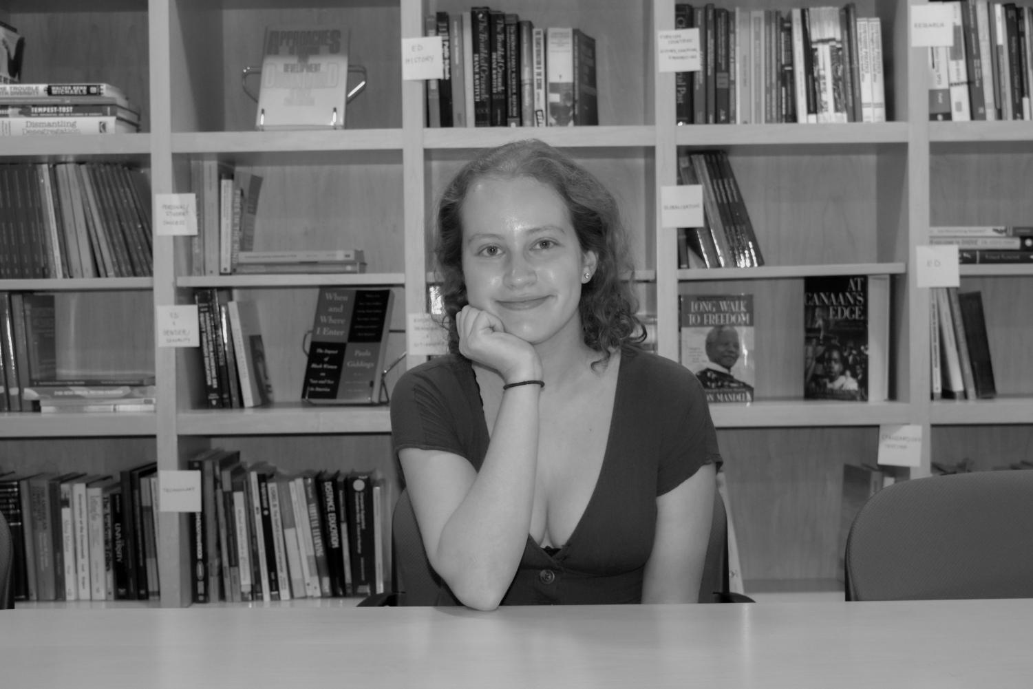 A black-and-white image of a female college student with light curly hair, a dark top, and a hair tie on her wrist sitting in a chair. She smiles at the camera with her chin resting in her hand in front of a bookshelf with four rows and five columns.