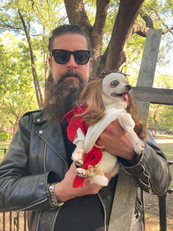 a middle-aged man wearing a leather jacket and a pair of black sunglasses holds a chihuahua in a brown wig and red costume.