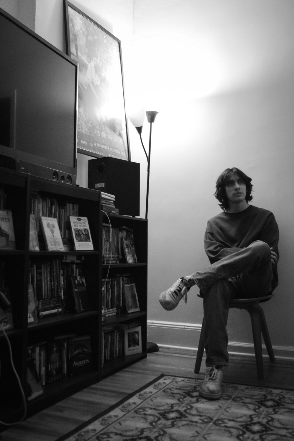 A black-and-white image of a male college student with long, fluffy, light hair wearing a plain crew neck sweatshirt, jeans, and light sneakers sitting in a chair next to a bookshelf. The bookshelf has a television on top and a full-length lamp next to it. The student has one leg crossed and is looking to their right.