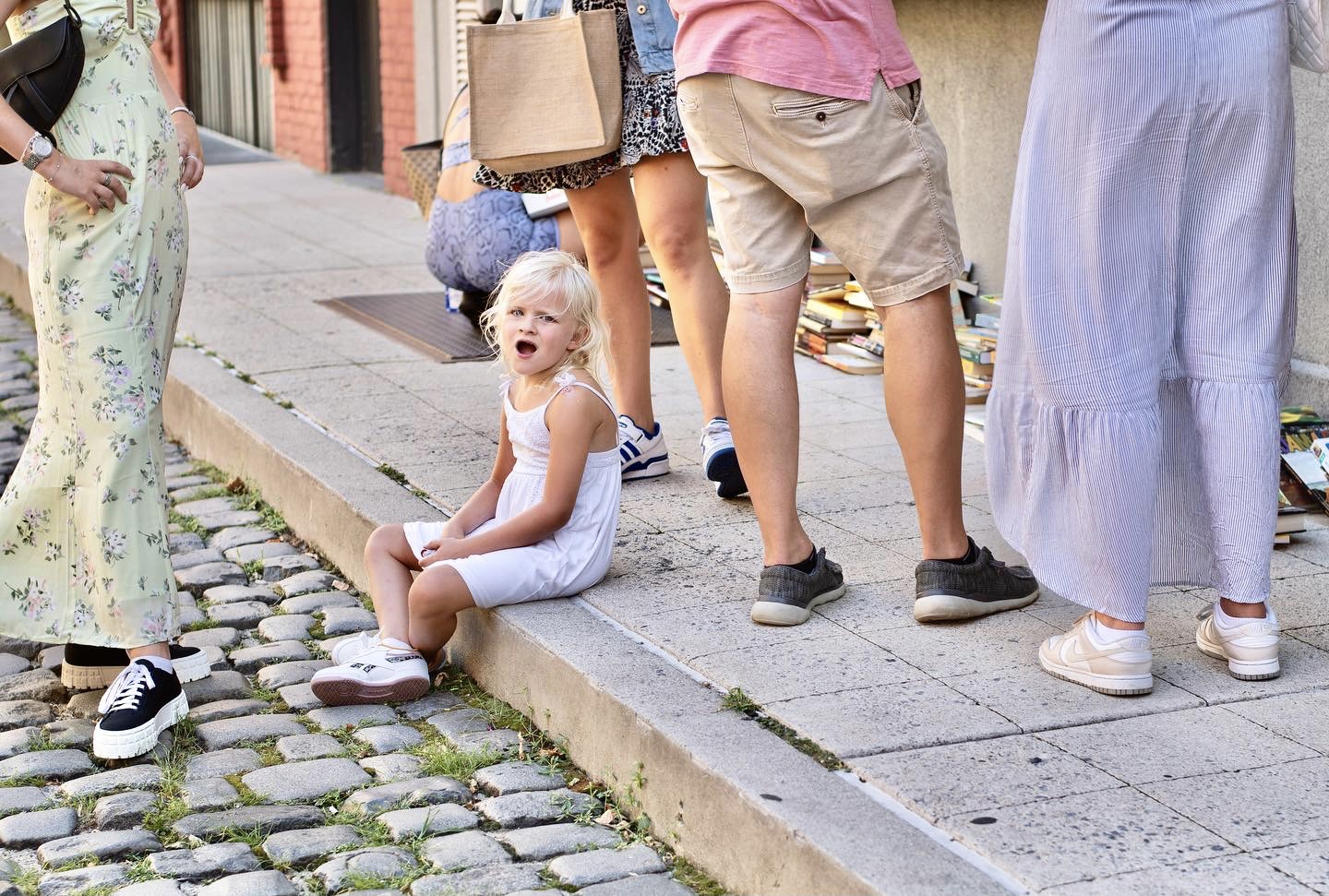 A little girl with blonde hair wears a sleeveless white dress and sits on a concrete sidewalk with her legs crossed.