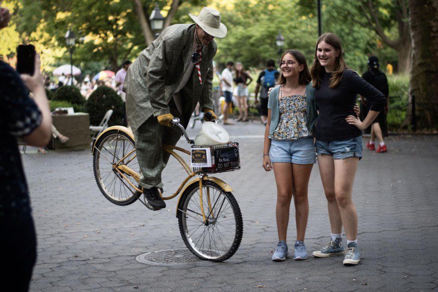 A man wearing a gray suit and a khaki hat does tricks on a bicycle as two girls both wearing jean shorts — one with a black top, and the other with a floral camisole and teal cardigan — pose for a photo together.