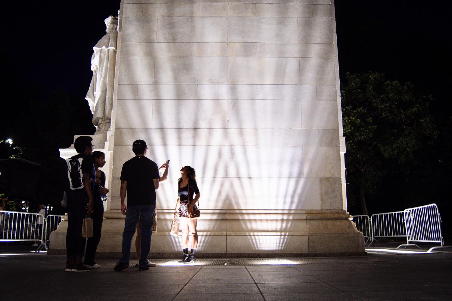 A woman stands alongside the Washington Square Arch at night. She is wearing a long-sleeved black top and brown shorts.