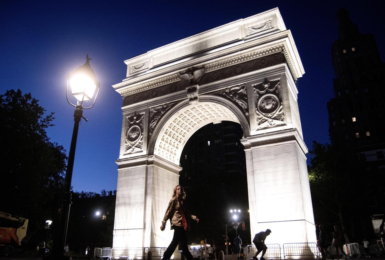 The marble Washington Square Arch illuminated by lights at night. To the left, a black lamp post, and at the bottom of the arch, a woman walking while wearing a brown long-sleeved shirt and black pants.