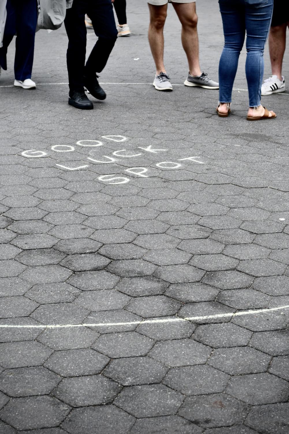 On concrete, a yellow chalk drawing of a circle that reads “Good Luck Spot” inside. The feet of people who are standing in the circle are visible.