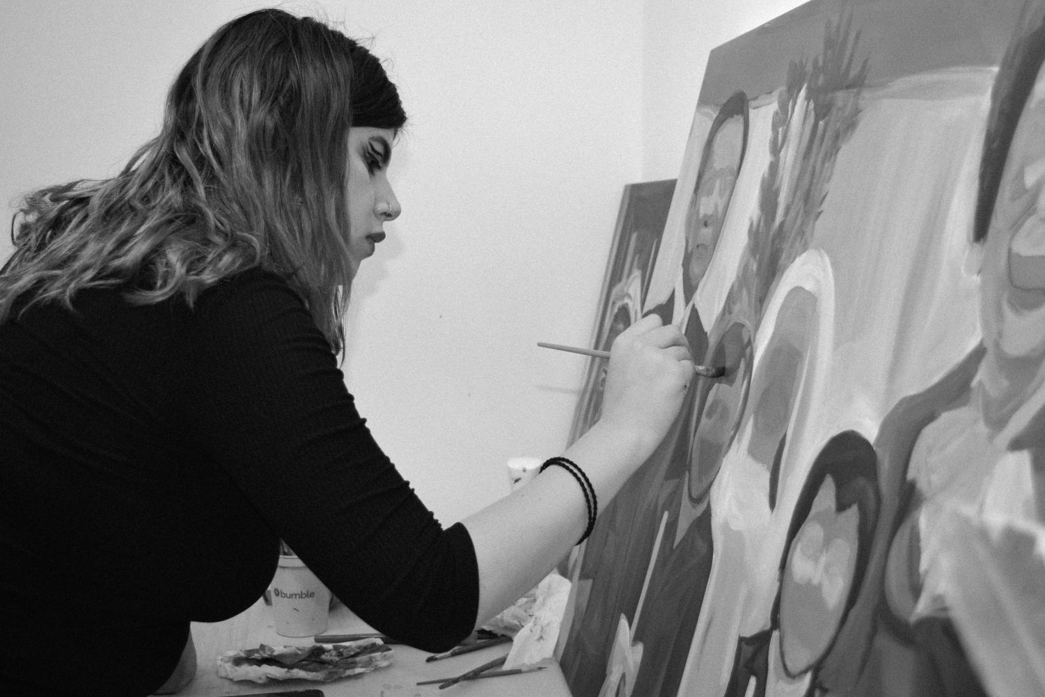 A black-and-white image of a female college student with medium-length, slightly curly hair, a dark long-sleeved top, and hair ties on her right wrist. She is holding up a paintbrush in her right hand to a canvas with an in-progress painting of multiple faceless figures.