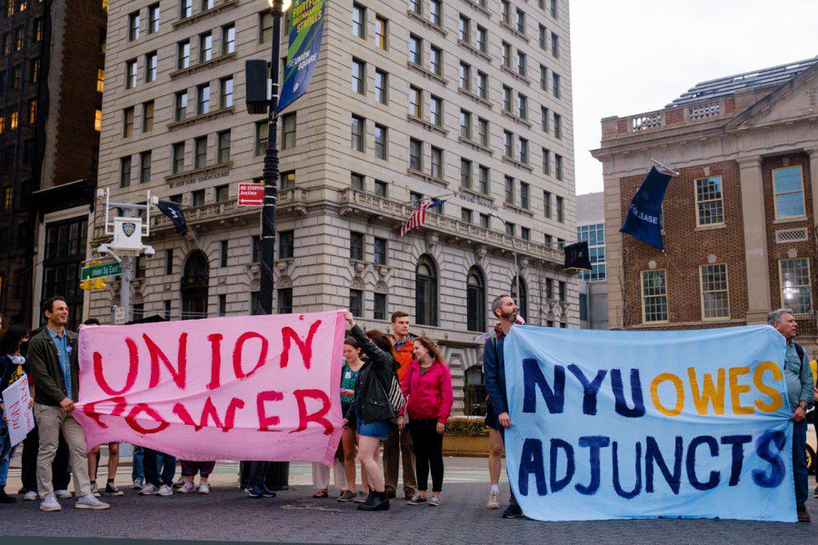 Protesters walk down University Place holding a large sign that reads “N.Y.U. owes adjuncts.” A police officer on a moped directs traffic in front of the marching protesters.