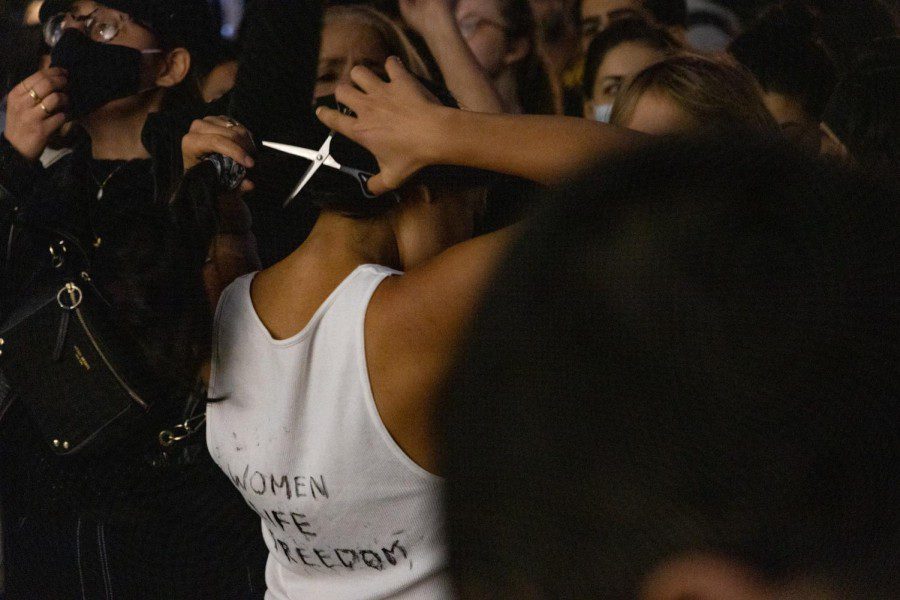 A female protester wearing a white shirt with text “WOMEN LIFE FREDDOM” cuts off her dark hair with a scissor.