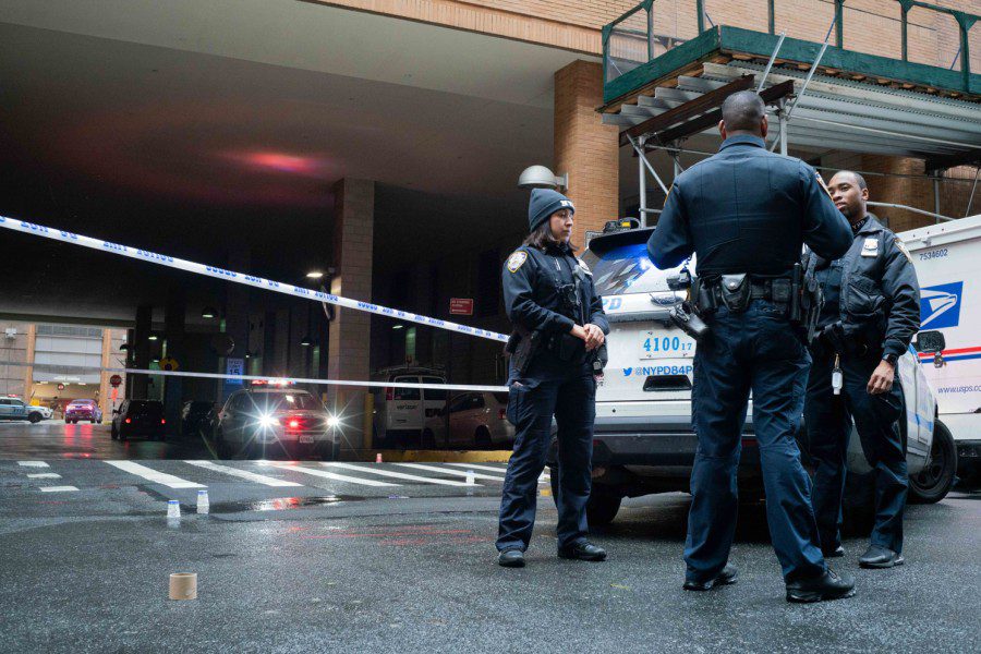 Three N.Y.P.D. officers stand next to a police vehicle. On the ground are two separate bullet markings. There is barricade tape in the background.