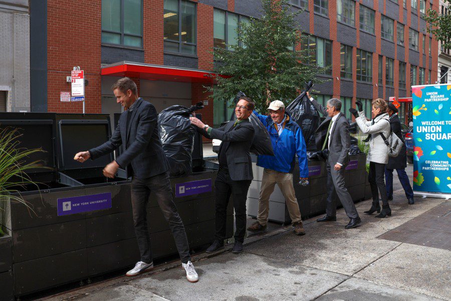 A line of people throw bags of trash into black garbage bins labeled “New York University.”