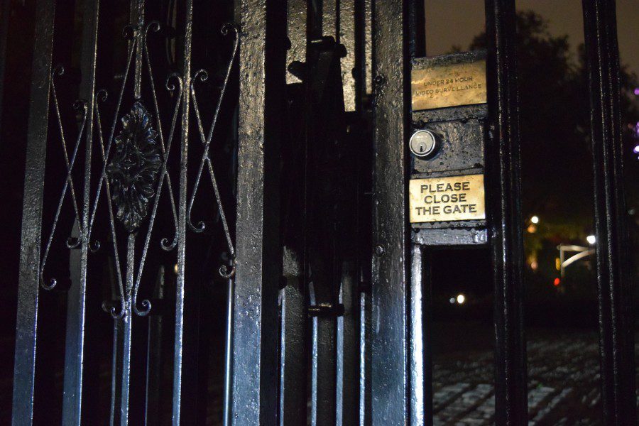 A nighttime photograph of a black, locked gate labeled “UNDER 24 HOUR VIDEO SURVEILLANCE” on the top and “PLEASE CLOSE THE GATE” on the bottom. Between the two gold-colored signs is a silver-colored lock.