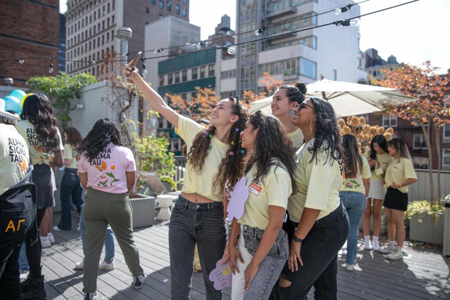 A group of four people wearing the same yellow sorority t-shirts all take a selfie with a smartphone. They stand on a rooftop with other sorority members in the background.