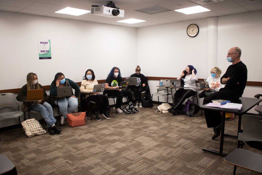 Seven students wear masks while sitting at their desks in an NYU classroom. A professor sitting on a table listens to a student.
