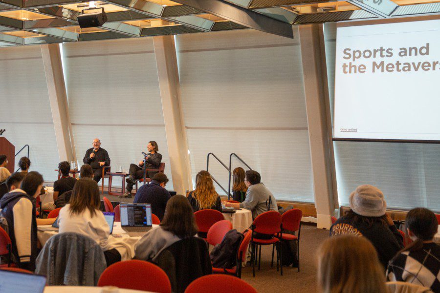 Two speakers sit on a stage, in front of an audience with a projector screen displaying “Sports and the Metaverse” against a white background.