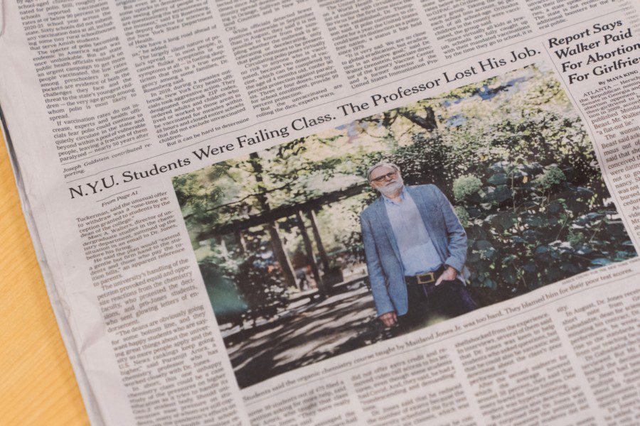 A photograph of a printed edition of The New York Times. The newspaper is turned to a page on which an article titled “N.Y.U. Students Were Failing Class. The Professor Lost His Job” is visible. In the newspaper, the article is accompanied by a photo of a male wearing a blue suit jacket standing in a park.