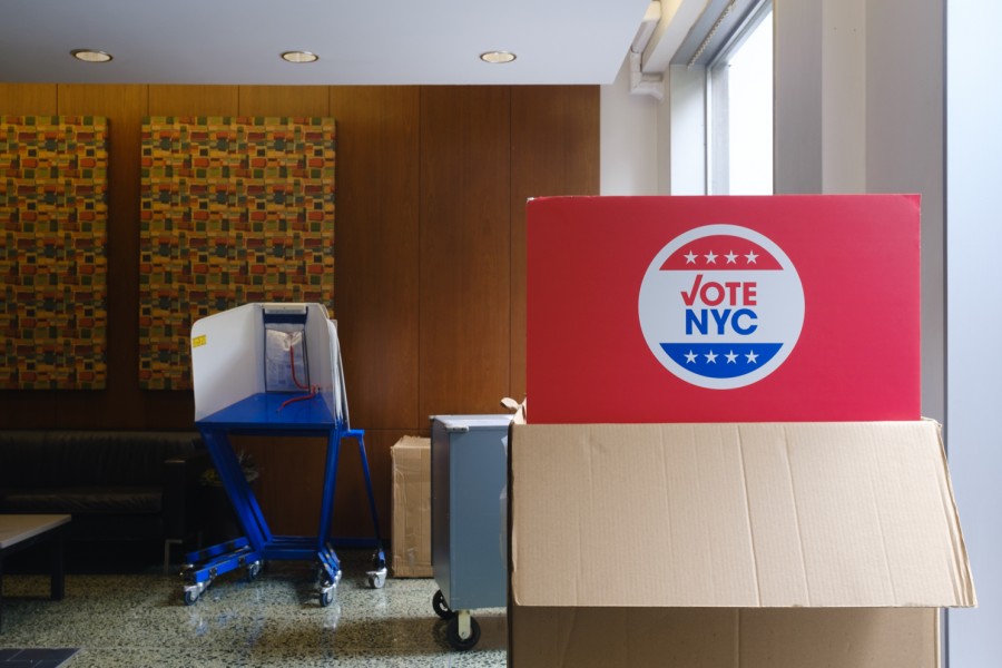 Inside a New York City polling site. A red "Vote N.Y.C." sign in the foreground, and a polling booth in the background.