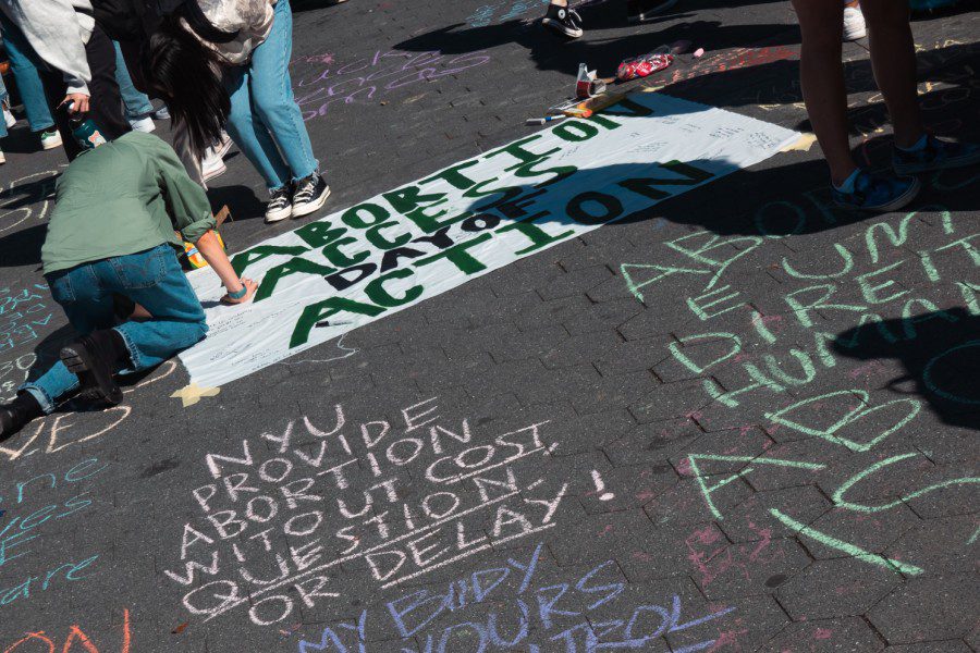 Two people writing on a poster laid on the ground with text “ABORTION ACCESS DAY OF ACTION.” Around the poster are texts written on the ground with chalk, one block of text says “NYU PROVIDE ABORTION WITHOUT COST, QUESTION, OR DELAY!”