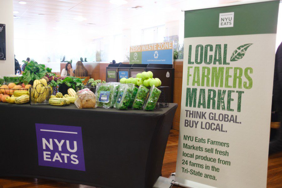 Photograph of a banner saying “N.Y.U. Farmer’s Market” on the right, and an assortment of fruits and vegetables on a table covered in a purple N.Y.U.-branded tablecloth on the left.