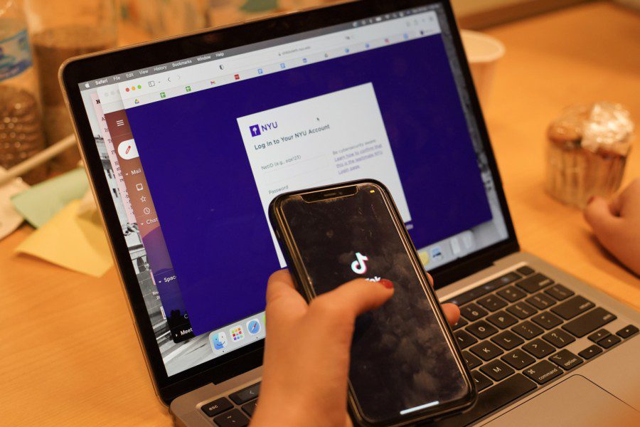 A hand holding a smartphone displaying TikTok’s splash screen. In the background is a laptop with N.Y.U.’s log-in webpage open.