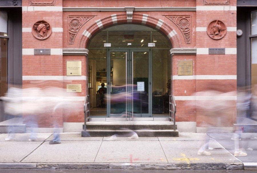 A group of people walking past the entrance to the building of 80 Washington Square East galleries with red bricks on the facade. The people are blurry because of a motion blur effect.