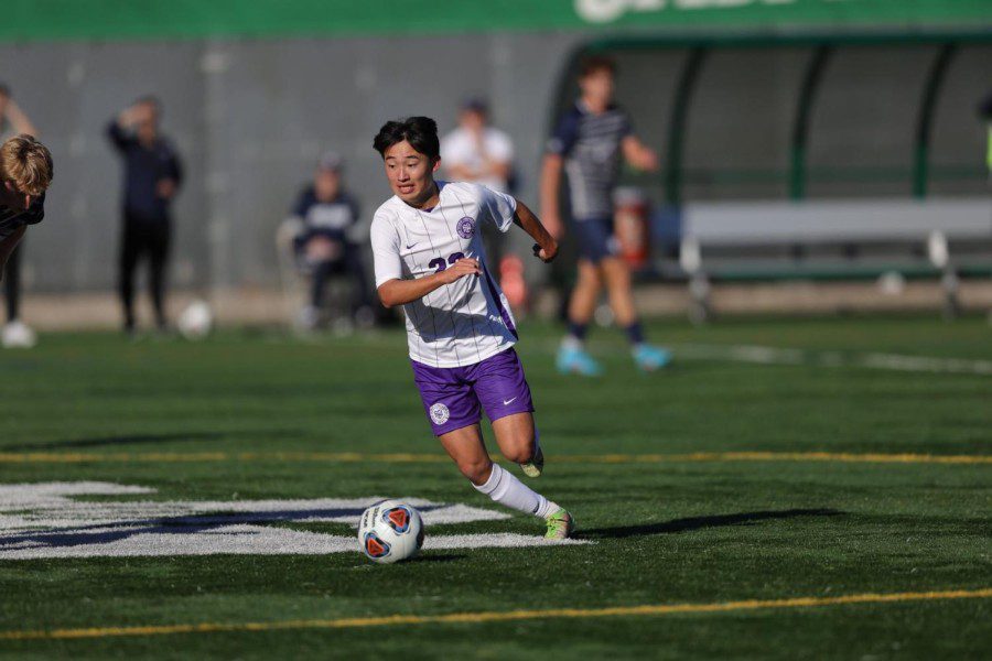Terry Nagai wears a white jersey, a pair of purple shorts is about to kick a soccer ball down the field. Opponents from Wilke University dressed in black are in the background.