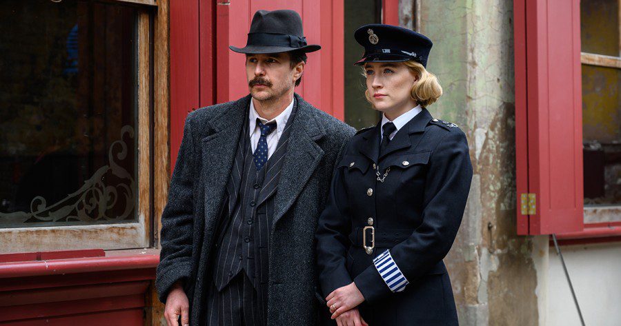 Actress Saoirse Ronan wears a police officer uniform and actor Sam Rockwell wears a suit, coat and hat as they both stand on the street in a scene from the film “See How They Run.”