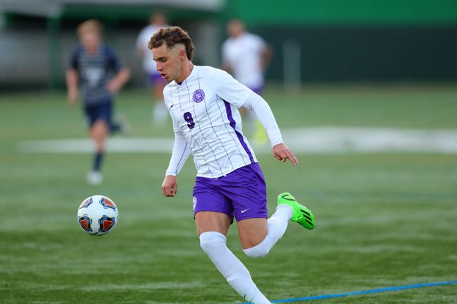 N.Y.U men’s soccer player Arkan Tahsildaroglu wears a white and purple jersey, a pair of purple shorts and neon green shoes as he prepares to kick a soccer ball on the field.