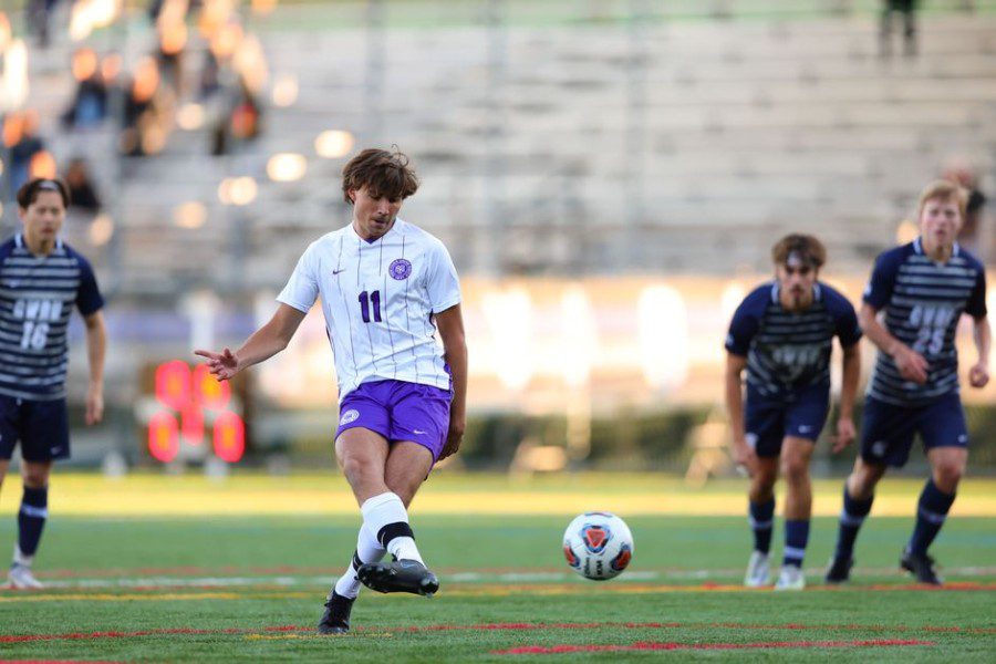 Bryce+Lexow%2C+wearing+a+purple+and+white+striped+NYU+jersey%2C+kicks+a+soccer+ball.+Three+opponents+wearing+black+jerseys+are+moving+in+the+background.