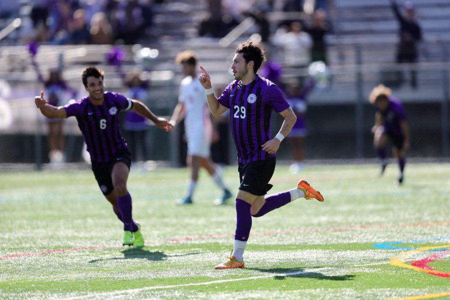 Alex Rovirosa-Illa, player number, and Joe Leslie, player number 29, run down the field in purple jerseys, black shorts and long socks. Rovirosa-Illa is wearing a pair of neon green soccer cleats. Leslie is wearing a pair of orange soccer cleats. They point forward with their index fingers. Blurred out in the background is a cheerleading team. A player from the opposing team wearing white jerseys and shorts and the.